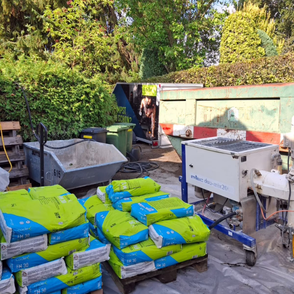 Bags of cement are stacked on pallets outdoors near a concrete mixer and a skip bin, surrounded by trees and bushes on a sunny day.