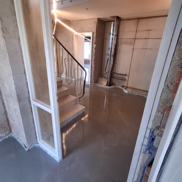 A room under renovation with smooth, wet cement flooring, unfinished walls, exposed pipes, and a staircase with a metal railing leading upstairs.