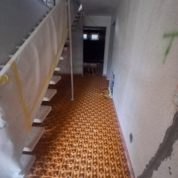 A hallway with stairs on the left side covered in protective material, and a floor covered with orange waffle-patterned underlayment panels. The walls are plain and lightly marked.