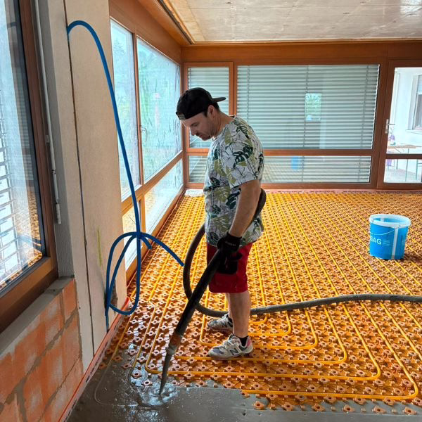 A person in casual clothes and a backwards cap pours cement over orange floor heating coils in a sunlit room under construction.