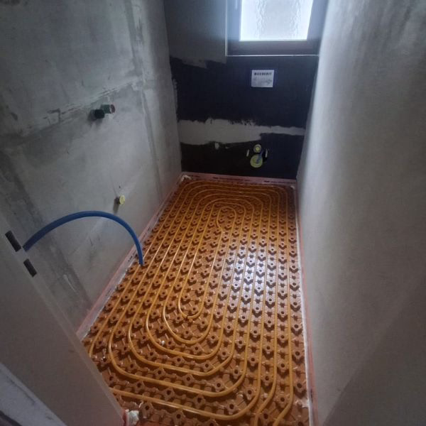 A small unfinished room with underfloor heating pipes installed in a grid pattern on the floor, surrounded by bare walls and a window at the far end.