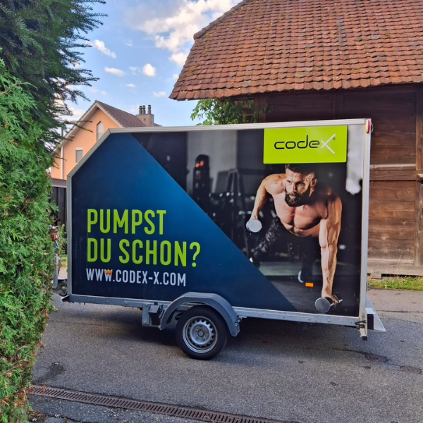 A trailer with an ad showing a muscular man doing push-ups with dumbbells, the text Pumpst du schon? and www.codex-x.com appears, promoting fitness. The trailer is parked on a residential street.
