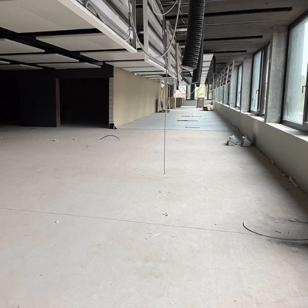 Large, empty room under construction with unfinished ceiling, exposed ducts, scattered debris, and large windows on the right letting in natural light.