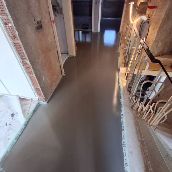 A hallway under renovation with freshly poured, smooth wet cement on the floor, tape along the walls, exposed wiring, and unfinished walls visible.