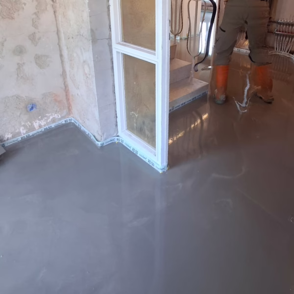 A person wearing orange boots stands on a staircase while freshly poured self-leveling cement covers the floor in a room under renovation.