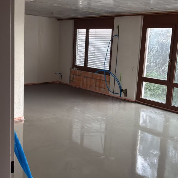 An empty room with large windows and a newly poured, shiny concrete floor. Blue pipes are visible on the wall, and bricks cover part of the lower wall, indicating ongoing renovation work.