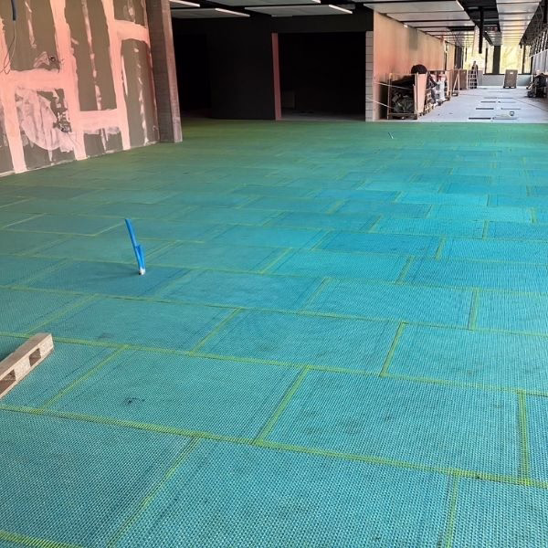 A large indoor room under construction with blue-green matting covering the floor, unfinished walls, tools, and construction materials visible in the background.