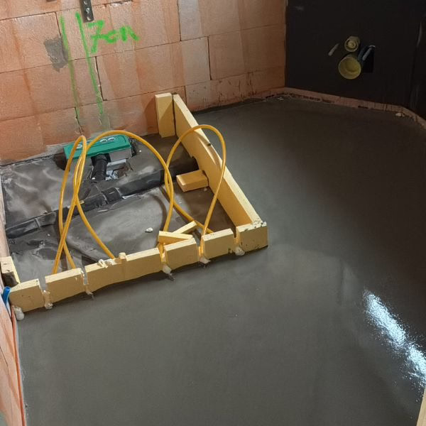 A bathroom under construction with wet concrete flooring, wooden forms, exposed pipes, and orange-tiled walls. Yellow tubing and unfinished plumbing are visible.