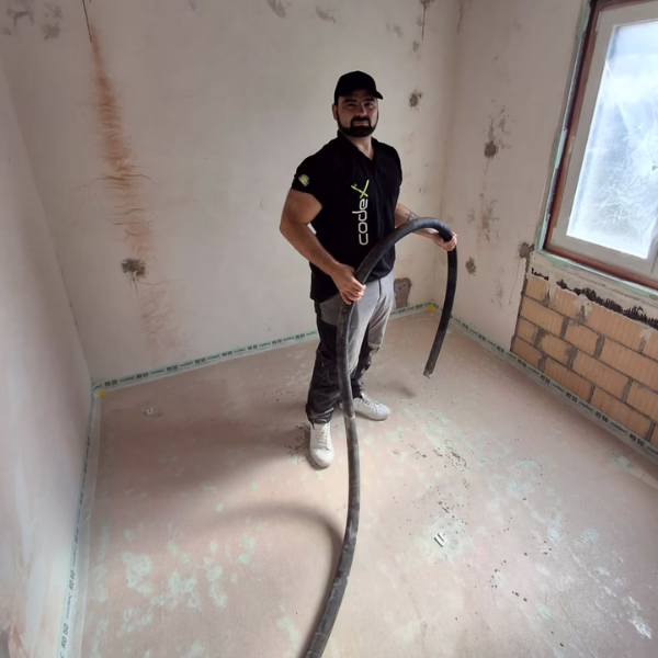 A man in a black shirt, pants, and cap stands in a partially renovated room holding a large black hose. The walls are unfinished, and the window is partially covered.