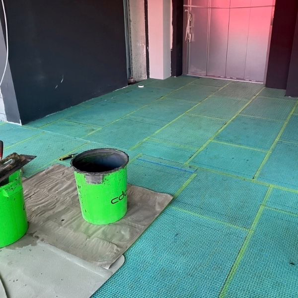 Two green buckets and a trowel rest on cardboard placed over a blue mesh underlayment on a floor, possibly in preparation for tiling. The surrounding walls are unfinished, and theres a pink-lit doorway in the background.