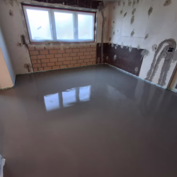 A room under renovation with unfinished walls and a freshly poured, smooth gray floor that reflects the window above a wall of exposed bricks.