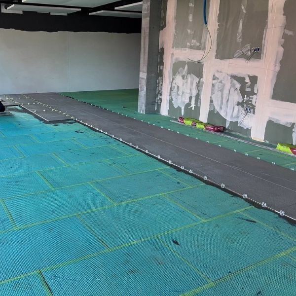 A room under construction with green subflooring, exposed wiring in unfinished drywall, and a long gray underlayment strip laid out, possibly for new flooring installation.