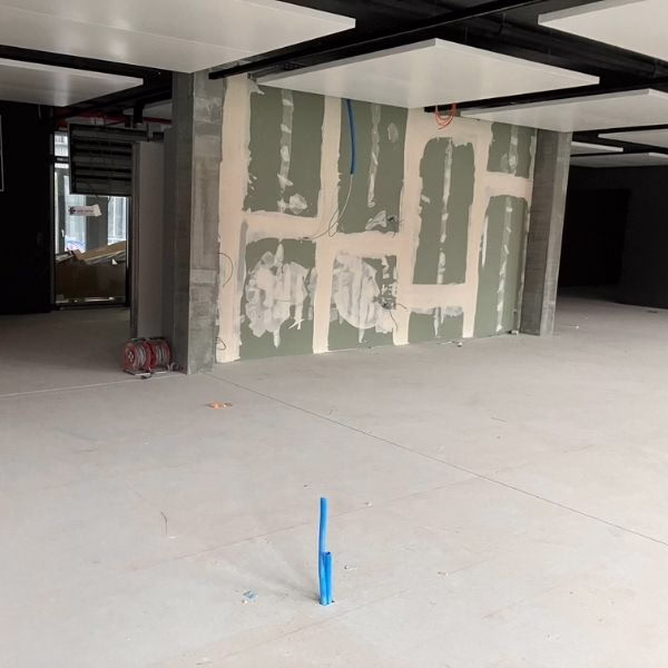 An unfinished room with exposed drywall and visible joint compound on the walls. The floor is bare, with a blue pipe protruding from it, and construction materials are visible in the background.