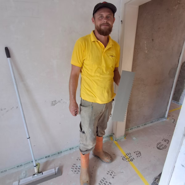 A man in a yellow shirt, cap, and orange boots stands in a partially renovated room, holding a notched trowel. A broom leans against the wall beside him. Floor shows footprints and some construction materials.