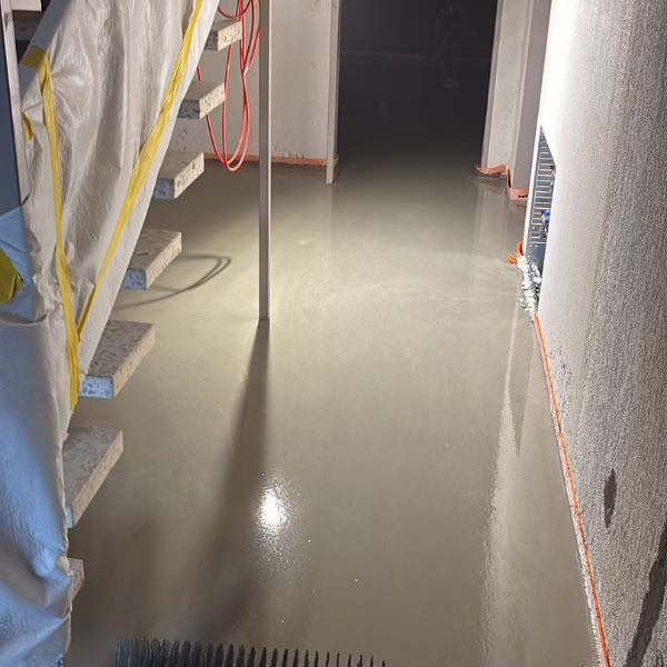 Freshly poured concrete floor in a building under construction, with stairs on the left, a broom in the foreground, and walls covered in protective plastic. The surface is smooth and reflective.