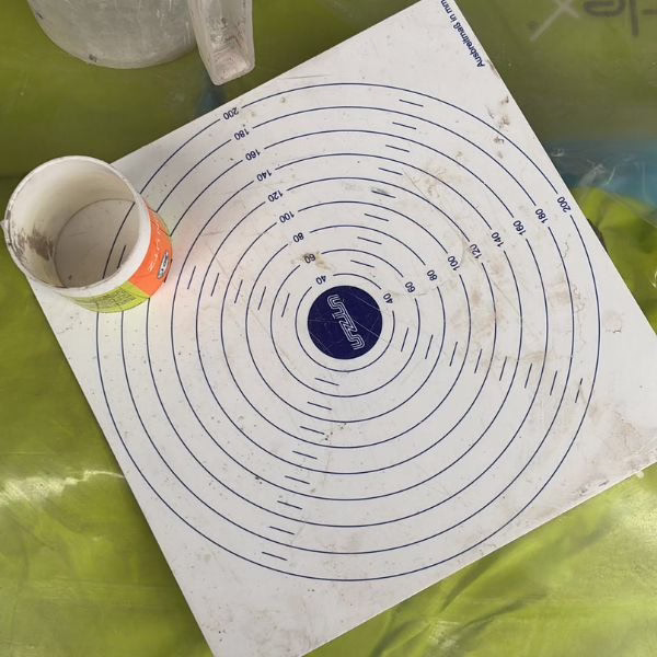 A dirty white circular target mat with blue concentric rings and numbers is placed on a green surface. A white paper cup lies on its side on top of the mat, partially covering some of the rings.