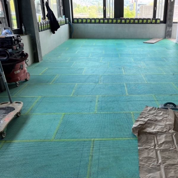 A large indoor space with green grid-patterned mats on the floor, surrounded by windows. Construction equipment and a piece of brown tarp are visible on the sides.