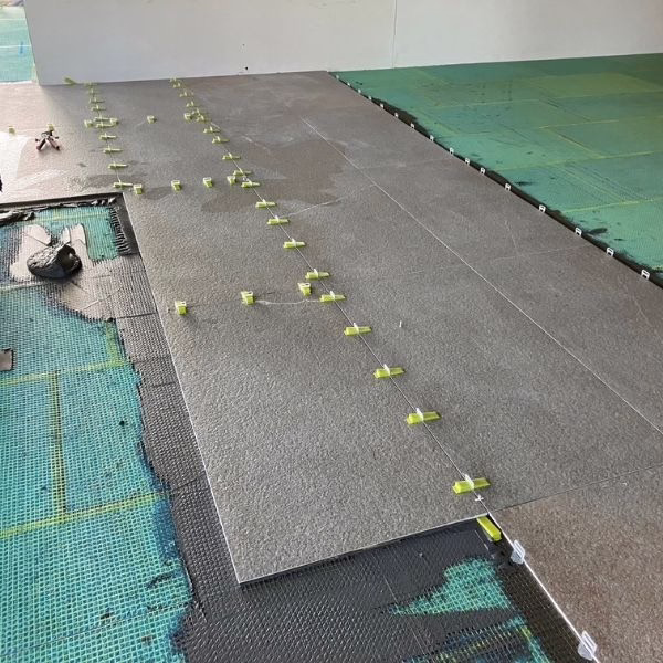 Large gray tiles are being installed on a green underlayment. Tile spacers and leveling clips are visible, and some tiles are in place while others are yet to be set. Tools are scattered nearby.