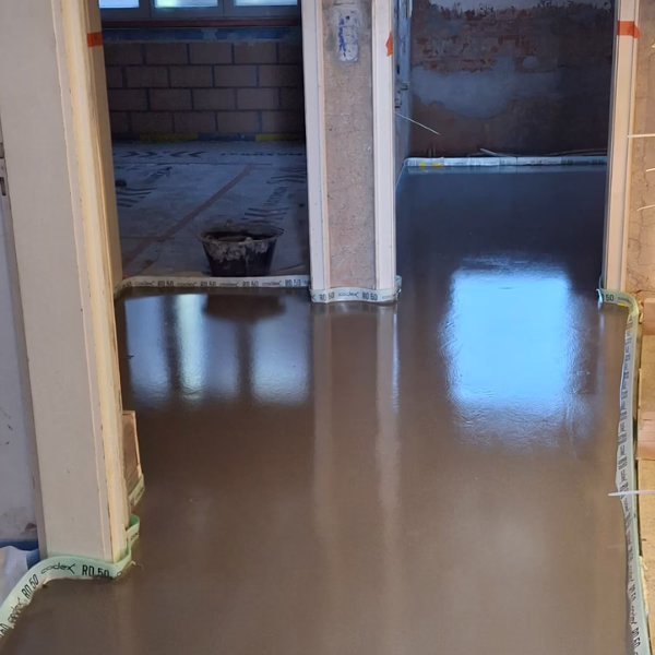 A freshly poured, smooth, wet concrete floor is curing inside a building under construction. Walls are unfinished, and construction materials are visible in the background.