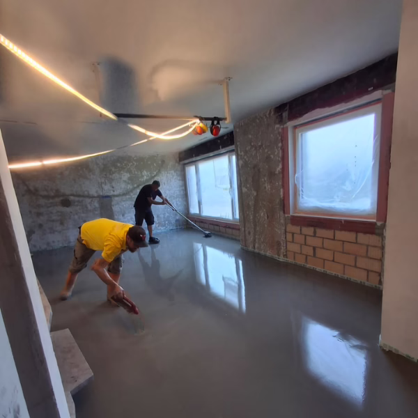 Two workers smooth freshly poured concrete on a floor inside a room under renovation, with large windows covered for protection and exposed brick visible on one wall.
