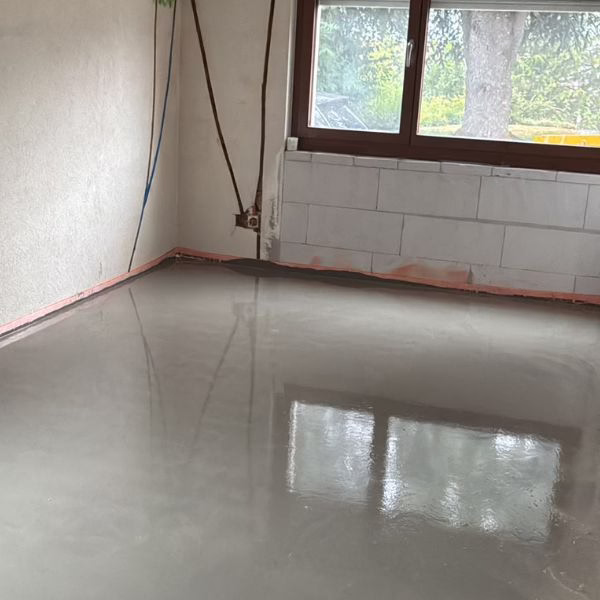 A room with unfinished walls and a large window, showing a freshly poured, smooth concrete floor that is still wet and reflective.