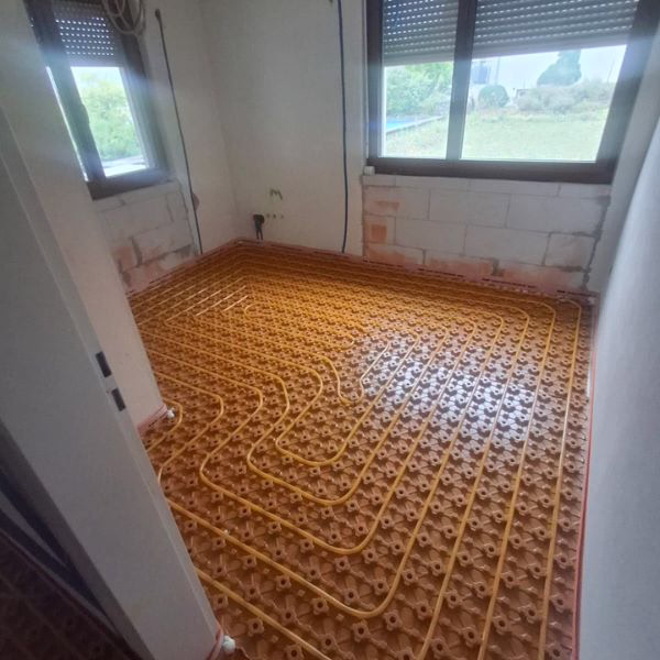 Room with underfloor heating pipes installed in a pattern on a yellow grid, unfinished walls, and large windows showing greenery outside.